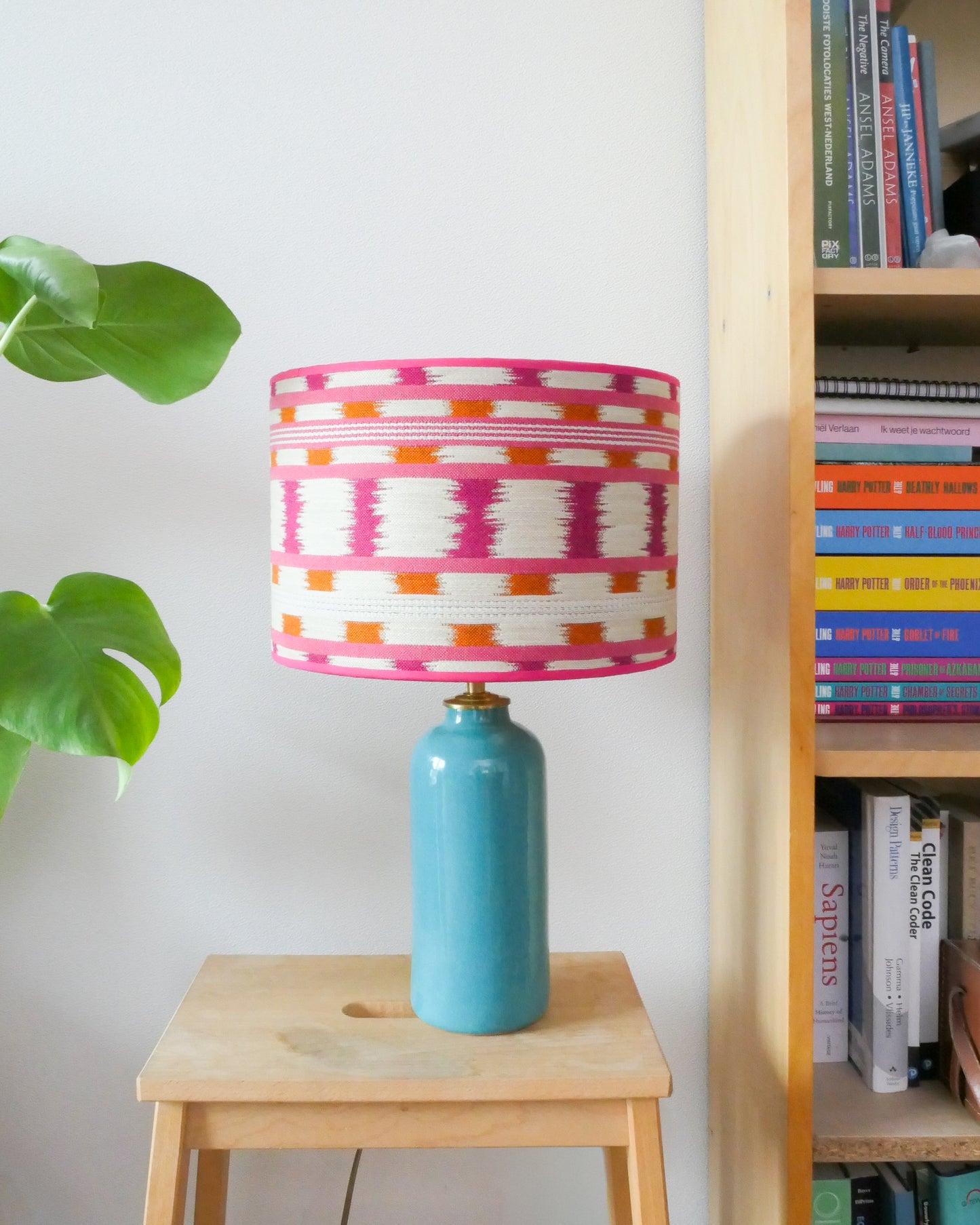 Tafellamp met een wit/roze lampenkap en een turquoise lampenvoet op een krukje naast een boekenkast.