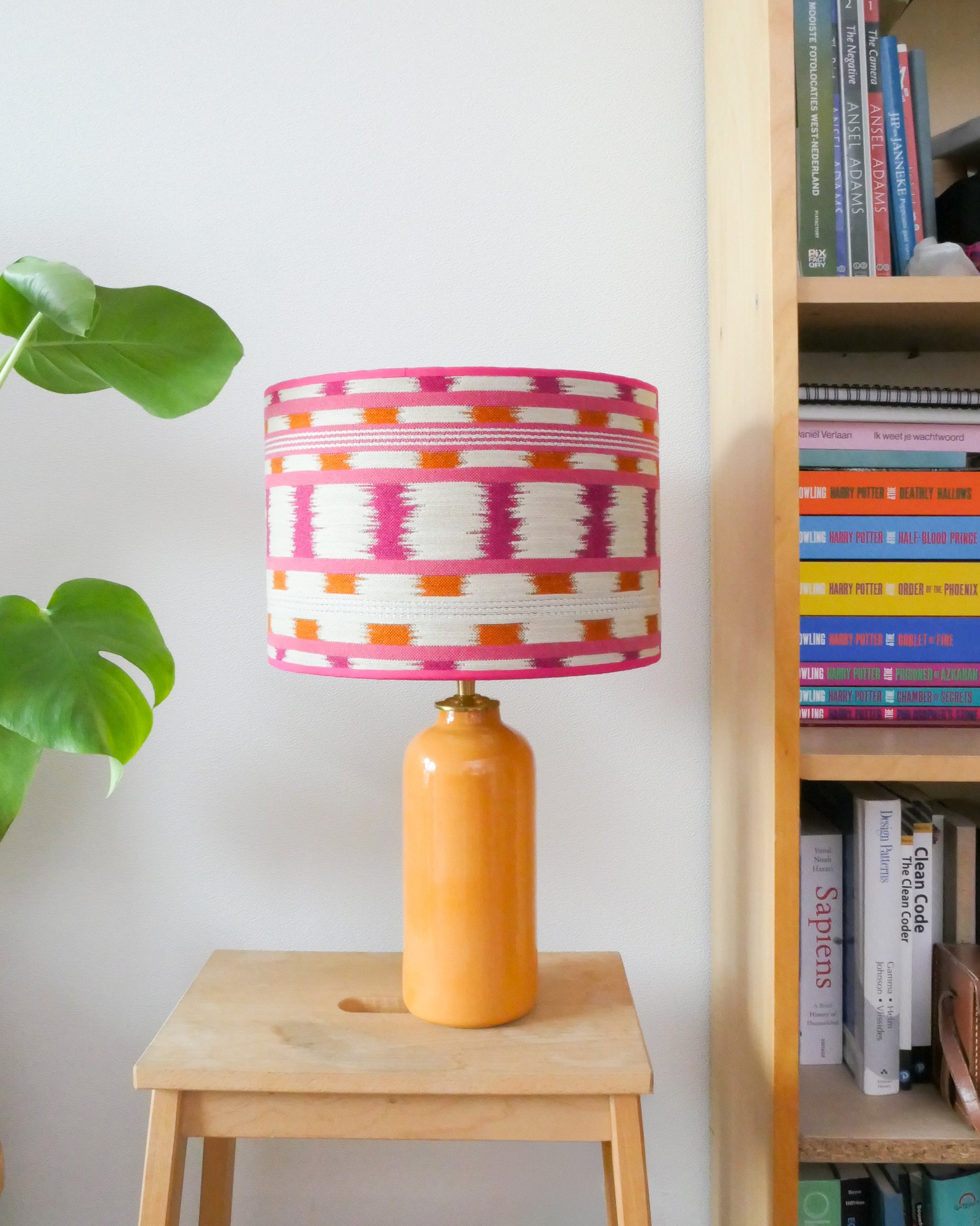 Tafellamp met wit/roze lampenkap en gele lampenvoet op een krukje naast een boekenkast.