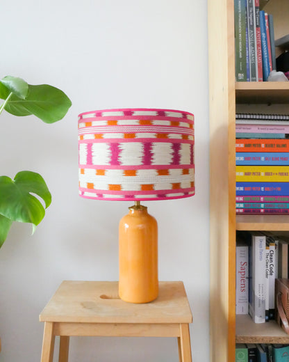 Tafellamp met wit/roze lampenkap en gele lampenvoet op een krukje naast een boekenkast.