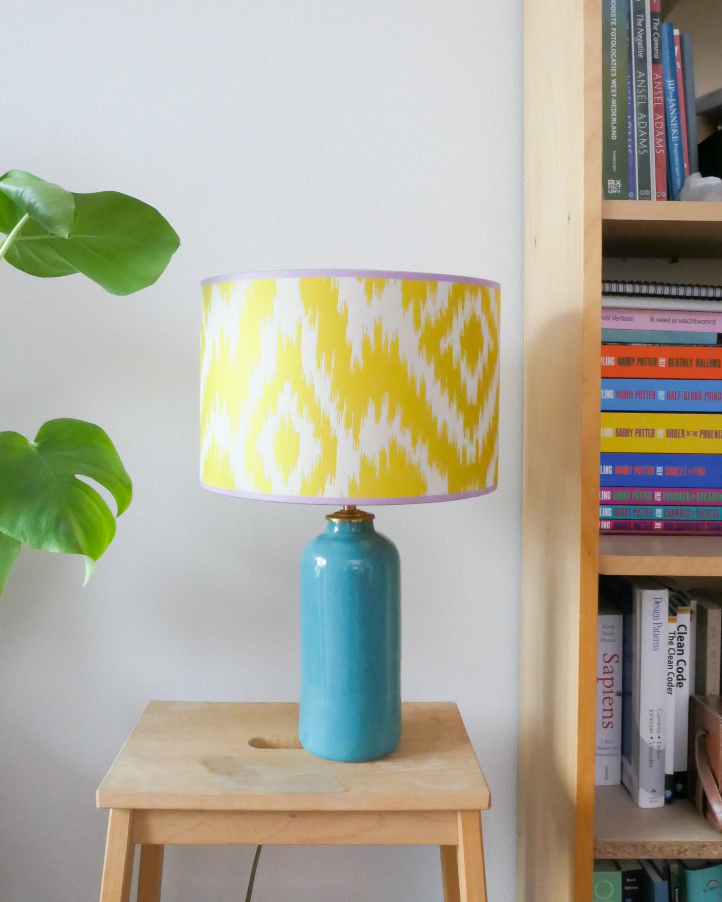 Tafellamp met een gele lampenkap en een turquoise lampenvoet op een krukje naast een boekenkast.