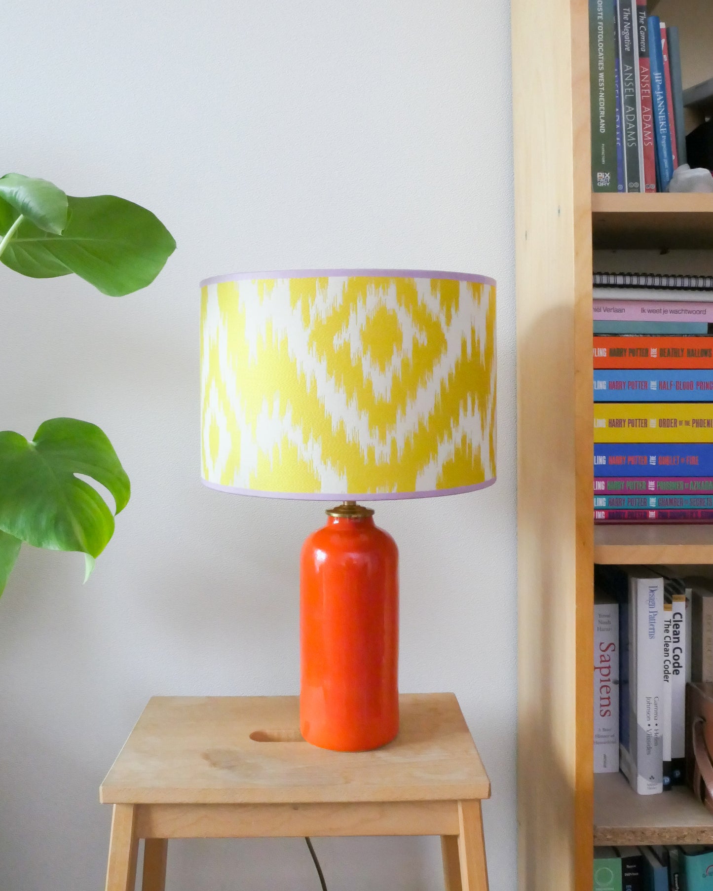 Tafellamp met een gele lampenkap en een knal oranje lampenvoet op een krukje naast een boekenkast.