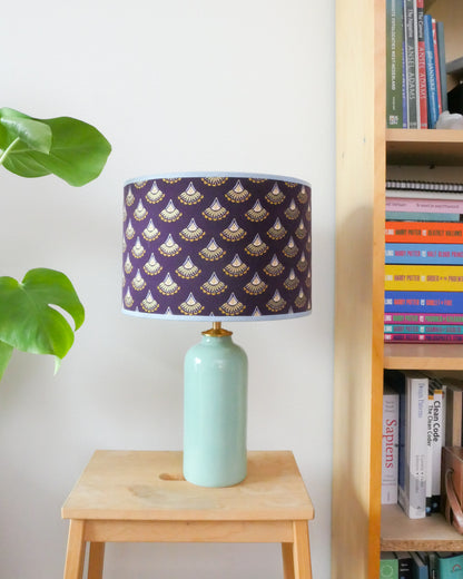 Tafellamp met een paarse lampenkap en een mint groene lampenvoet op een krukje naast een boekenkast.