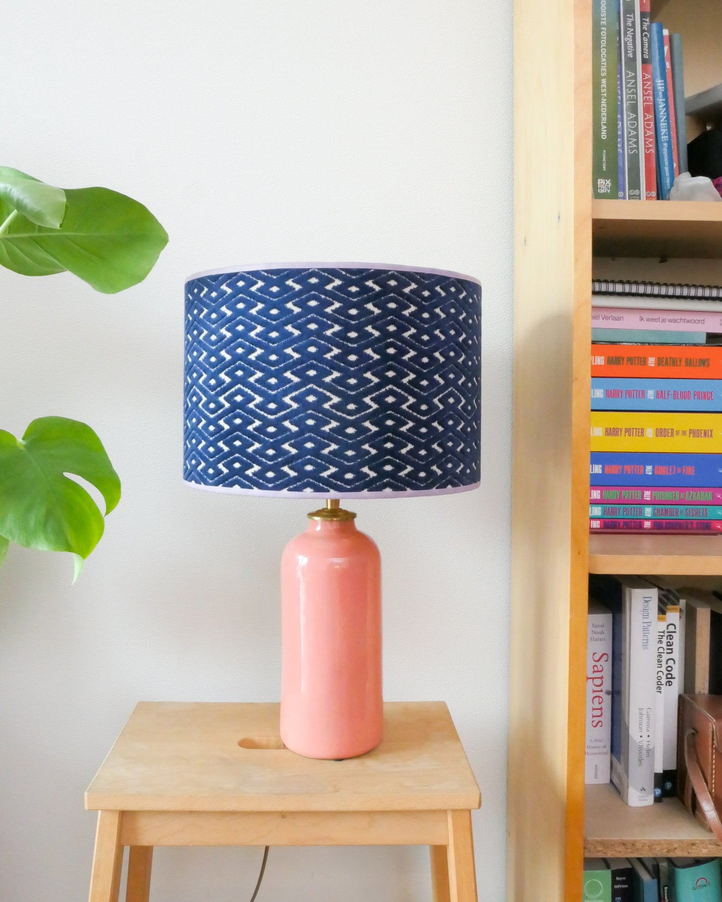 Tafellamp met blauwe lampenkap en licht roze lampenvoet op een krukje naast een boekenkast.