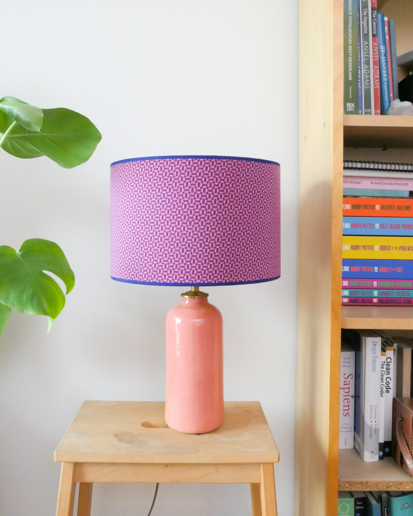 Tafellamp met een paars/roze lampenkap en een licht roze lampenvoet op een krukje naast een boekenkast.