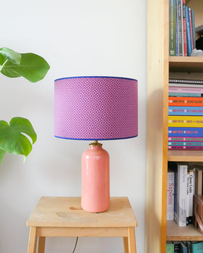 Tafellamp met een paars/roze lampenkap en een licht roze lampenvoet op een krukje naast een boekenkast.